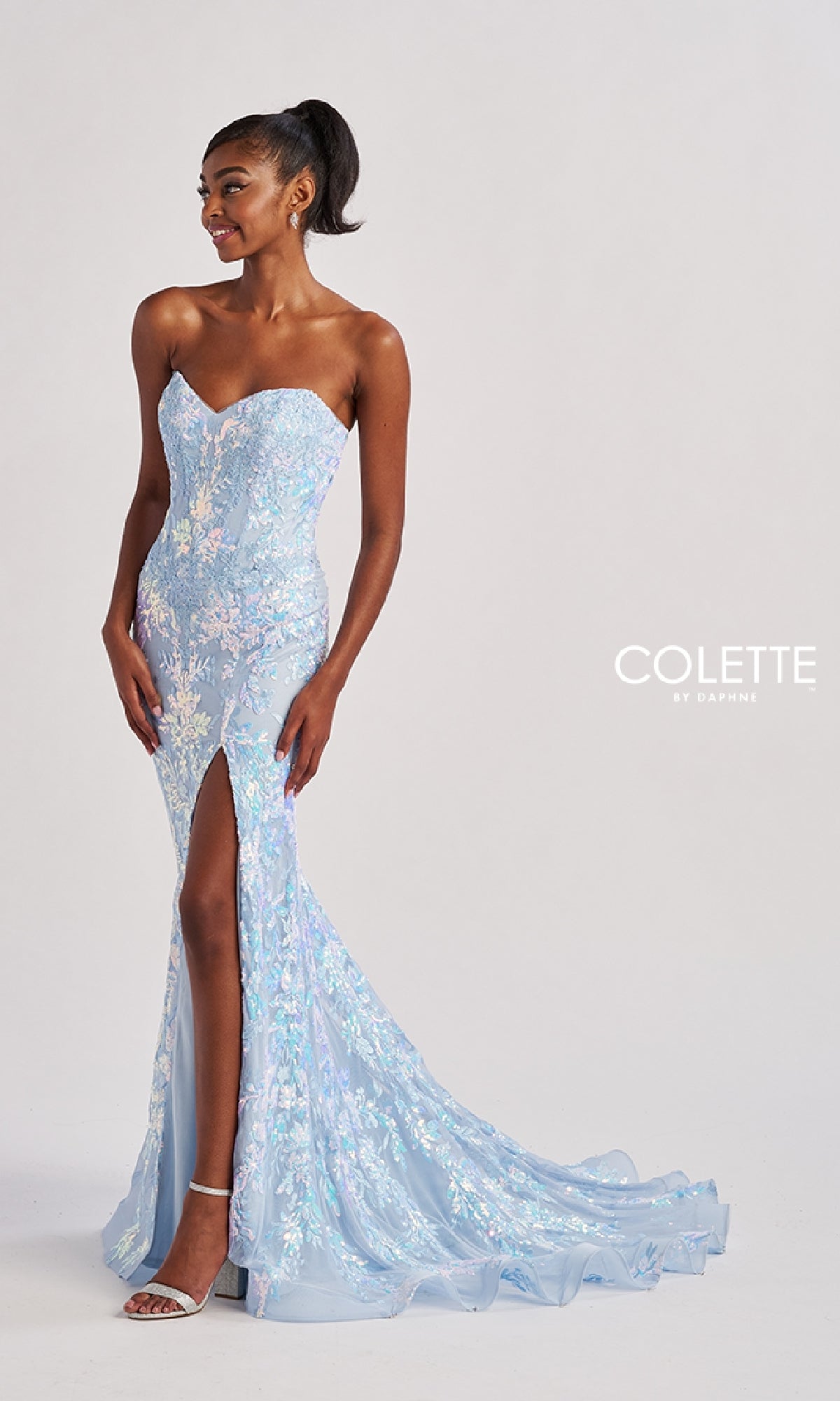 A woman wears the Colette CL8440 Long Prom Dress—a strapless, light blue gown with floral lace embroidery, floor-length skirt, thigh-high slit, and boned corset bodice—standing out against a plain background.